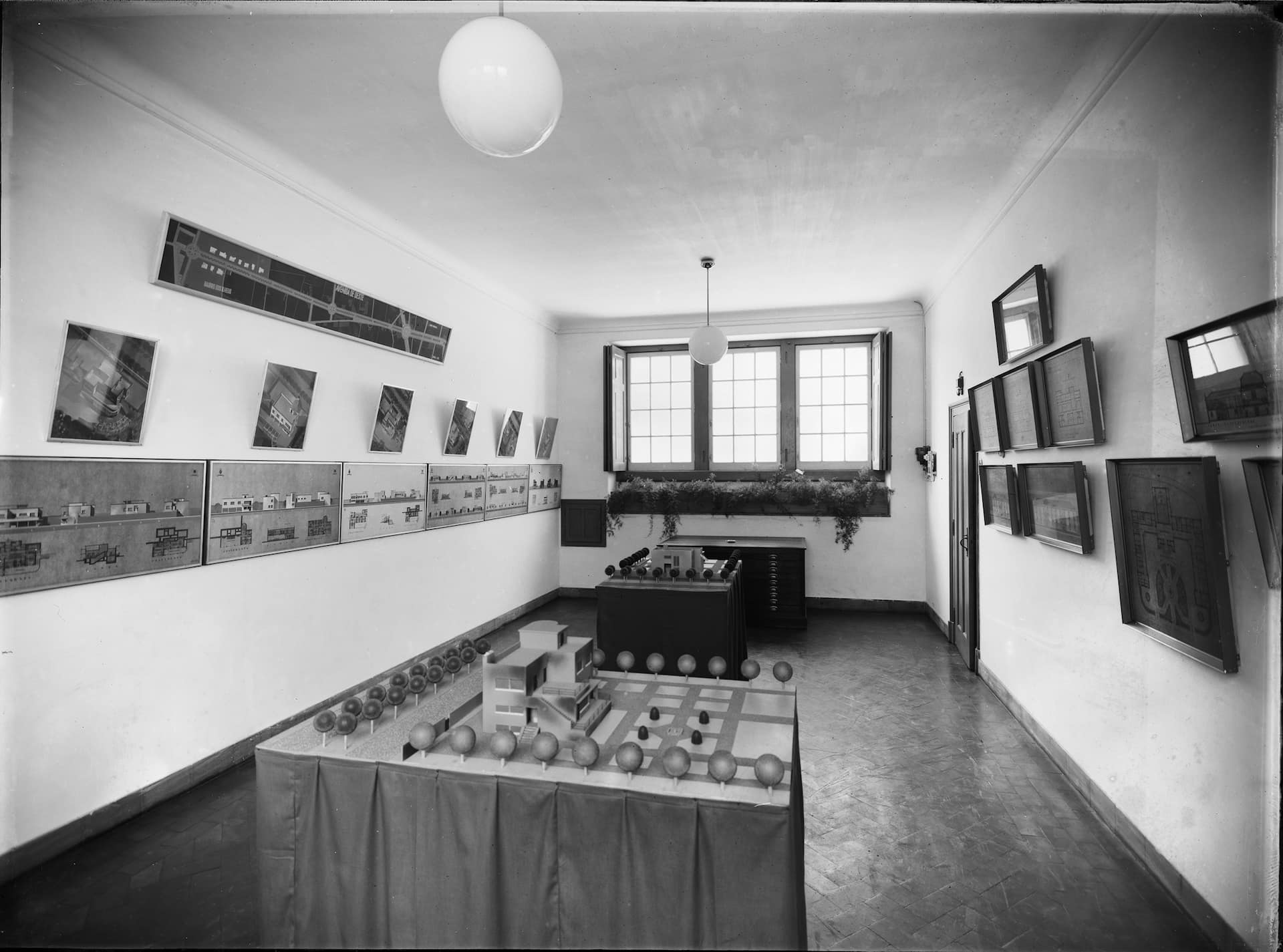 Exposição dos Melhoramentos "da Cidade do Funchal". Maquetas e técnica mista sobre papel colado sobre contraplacado. Atelier de Carlos Ramos (1897-1969), Lisboa, dezembro de 1931. Fotografia do estúdio de Mário Novais (1899-1967). ABM/ARM, CFT003.101688.ia, Funchal, ilha da Madeira. O arquiteto João Carlos Chambers de Oliveira Ramos (1897-1969), natural do Porto, formou-se pela Escola de Belas-Artes de Lisboa, dado o pai, Manuel Maria de Oliveira Ramos (1862-1931), ter sido convidado para lecionar nessa cidade a cátedra de História na Faculdade de Letras. Cresceu, assim, num privilegiado ambiente intelectual, tendo privado, inclusivamente, com o arquiteto Ventura Terra (1866-1919), o que o levou, em 1915, a fazer exame de admissão ao curso especial de arquitetura civil. Teria sido nesse quadro que, em 1931, a Câmara Municipal do Funchal e a Junta Geral do Distrito solicitaram a Carlos Ramos, uma nova proposta de urbanização para a cidade que, embora enraizada no plano inicial do seu mentor e mestre Ventura Terra, não o segue totalmente, acrescentando, inclusivamente, alguns elementos novos. Em março do seguinte ano de 1932, em conferência para o Palácio de São Lourenço, planeada em Lisboa, em dezembro anterior (RAMOS, Carlos, R 12-13 – Urbanização do Funchal, março 1932, policopiado), mas realizada a 5 desse mês de março, no Teatro Municipal ( DN , F., 5 mar. 1932, p. 1), explica, precisamente, que o seu trabalho não deveria ser considerado um “ plano de urbanização ”, que implicaria um outro trabalho mais maduro e alargado, com levantamentos gerais que não estavam feitos, mas somente um “ Plano de Melhoramentos ”. Mostrando já o caminho de pedagogo que abraçaria nos anos seguintes (1940), então na Escola de Arquitetura do Porto, que viria a dirigir (1952), não deixa de chamar a atenção para a necessidade de “ memórias descritivas ”, salvaguardando nessa altura, que essa chamada de atenção não incluía o vice-presidente da câmara, então o escultor Francisco Franco (1885-1955), que sempre assim fazia com os seus trabalhos. Carlos Ramos passara nos meados de 1931 algumas semanas no Funchal e, a 28 fev. 1932, apresentava numa exposição nas instalações da câmara do Funchal com as suas propostas, algumas em plantas, por certo coloridas e de grandes dimensões, nos finais do ano anterior já apresentadas em Lisboa e com algum destaque na imprensa da capital. Os elementos que chegaram aos nossos dias nos arquivos da CMF, essencialmente, são as fotografias então efetuadas em Lisboa pelo novo estúdio de Mário Novais (1899-1967), precisamente nesse ano de 1931, espólio hoje integrado na Biblioteca de Arte da Fundação Calouste Gulbenkian, mas somente a produção de 1933 a 1983, pela que a destes primeiros anos, só muito pontualmente ali consta, como o projeto revivalista para a câmara municipal de São Vicente, dado como de 1931, mas fotografia, provavelmente, de 1933. A exposição do Funchal foi fotografada por Manuel Perestrello (1893-1980), a 1 mar., tendo o DN publicado uma dessas imagens ( DN , 3 mar. 1932, p. 1), mas até ao momento não foi possível localizar os negativos. Como Carlos Ramos expôs na sua palestra do Funchal (Ramos, policopiado cit.), o trabalho dado como mais importante, que seria o de adaptação do antigo edifício da Misericórdia do Funchal a sede da Junta Geral, assunto que levantava então vasta polémica. A opção já ia para a reabilitação parcial da fachada para a Avenida Arriaga e a remontagem da restante para a nova Avenida Zarco, o que veio a acontecer alguns anos depois, estando em discussão entre jun. e jul. 1936, com destaque para o periódico Re-Nhau-Nhau , tendo sido efetuado em 1937 e o projeto depois do novo corpo, onde se encontrava o Circo Paris , entregue ao arquiteto Januário Godinho (1910-1990), em ago. 1945. O arquiteto abordou depois o projeto dos novos Paços do Concelho de São Vicente, de que parte do material chegou aos nossos dias, bem ao gosto revivalista do chamado “ Português Suave ”, que o mesmo arquiteto entendia como “ a arquitetura que caracteriza a maioria dos Paços do Concelho de Portugal, ou seja, a nossa arquitetura do século XVIII ”, o que é, no mínimo, uma meia verdade. Em 1940 existe outra versão, hoje no ABM e não deixa de ser estranho que quase na mesma data haja também projeto de 1939 do arquiteto Edmundo Tavares (1892-1983), único arquiteto a residir na Ilha a partir de 1932, mas ao gosto modernista e não revivalista, como esse e que se repete em 1942. Nenhum veio a ser levantado, já havendo projeto não identificado de cerca de 1900, também não levantado. Especial destaque foi dado em seguida aos projetos para os fontanários do Funchal, que Carlos Ramos organizou em 3 conjuntos, marcados “ com as armas da Câmara Municipal do Funchal e a data da sua construção ”, para “ em todos os tempos […] lembrar os nomes daqueles que tiveram essa generosa e singela lembrança ”. O “ de maiores proporções, destinado, sem dúvida, a ornamentar qualquer praça, largo, topo ou gaveto de maior importância ”, era concebido com 3 faces e independente, sendo os dois restantes tipos mais simples e destinados a ficarem encostados a paredes, todos dotados de painéis de azulejos. Parece somente se ter localizado o projeto para o primeiro, embora de 4 e não de 3 faces, datado de jun. 1933 e, depois, levantado no Largo Visconde do Ribeiro Real, mas sem as armas da cidade. Nos anos seguintes, entretanto, um pouco por toda a cidade do Funchal foram sendo levantados estes fontanários com a indicação de C.M.F. , a data e painéis de azulejos, projetados por Carlos Ramos. Longa memória descritiva foi apresentada sobre a construção dos Bairros Sociais, destinados a serem levantados pelas encostas da cidade, constituindo “ o grosso das habitações operárias e das classes médias ”, de que vieram vários desenhos e maquetas, reconhecíveis nas fotografias, os chamados “ bairros económicos ”, mas que parecem não ter chegado até nós. O mesmo se passou com o projeto do Sanatório, embora os desenhos tenham chegado aos nossos dias, mas não a maqueta e tendo o projeto de 1941, sido entregue depois, nas várias ampliações, ao arquiteto Vasco de Morais Palmeiro (Regaleira) (1897-1968), responsável por essa área na Assistência Nacional aos Tuberculosos (ANS). A nova avenida oeste de saída do centro da cidade, atual Av. do Infante, mereceu igualmente uma muito especial atenção, assim como o então chamado “ Bairro dos Ilhéus ”, previsto para bordear a área norte. O arquiteto tece uma série de considerações, de forma que as habitações unifamiliares e independentes a levantar ali obedecessem a uma única gramática arquitetónica “ à antiga portuguesa ”, a “ uma arquitetura tradicionalista, com os seus beirados, azulejos, rótulas, pináculos, vasos de flores, etc., etc .”. Aconselha, inclusivamente, a edilidade a criar “ um conselho técnico e de arte ” com um engenheiro e um arquiteto, visto tratar-se de uma via destinada às classes mais abastadas, devendo esta nova avenida constituir-se arquitetonicamente como uma via nobre e erudita. Igual cuidado apresentou para os projetos dos “ jardins, parques e miradouros ”, apresentando desenhos para o Jardim da Pontinha, Largo António Nobre, Largo Ribeiro Real, Largo da Bela Vista (Cruzes), Largo do Torreão, Praça de Tenerife, com o pormenor de seguir a ideia de Ventura Terra de cobrir a ribeira, que passaria a Avenida de João Gomes e o Largo do Socorro ou de Santa Maria Maior, com miradouro sobre a Praia da Barreirinha. O arquiteto Carlos Ramos foi alvo no Funchal de algumas homenagens, como a 7 mar., no restaurante-esplanada do Terreiro da Luta e pediu, entretanto, autorização às autoridades locais para apresentar os seus trabalhos em Londres, o que não sabemos se veio a acontecer, tendo embarcado no Carvalho Araújo , para Lisboa, a 10 desse mês de março ( DN , 10 mar. 1932, p. 1). Manteria, entretanto, contatos com o Funchal e a Madeira, sendo o principal responsável pela reabilitação da sede da Câmara Municipal do Funchal, em 1940, em parceria com o arquiteto Raul Lino (1879-1974), altura em que teria projetado alguns dos prédios da envolvência, como o das Finanças. E, inclusivamente, com a parceria que viria a montar com o filho, o arquiteto Carlos Manuel Oliveira Ramos (1922-2012), responsável, depois, pelo Hotel Hilton Madeira , encomendado então pela Lignum , nos finais de 60, obras visitadas a 16 jun. 1970 pelo almirante Américo Thomaz (1894-1987) e, inaugurado pelo mesmo, a 19 set. 1971, hotel depois denominado Madeira Palácio . Carlos Ramos (pai) faleceu no Porto, a 1 jul. 1969. Bibliografia: AAVV (1986), Carlos Ramos: exposição retrospetiva da sua obra . Lisboa: Fundação Calouste Gulbenkian (exposição itinerante); COUTINHO, Bárbara dos Santos (2001), Carlos Ramos (1897-1969): Obra, Pensamento e Acção. A Procura do Compromisso entre o Modernismo e a Tradição . Lisboa: FCSH/ UNL; FERNANDES, José Manuel (abr. 2014), Carlos Ramos, Arquiteturas do século XX em Portugal , Lisboa, IN/Casa da Moeda; RAMOS, Carlos (dez. 1931), “Urbanização do Funchal, conferência realizada no Palácio de S. Lourenço, março 1932” (policopiado, ARM, R 12-13); VASCONCELOS, Teresa (2008), O Plano Ventura Terra e a Modernização do Funchal (Primeira Metade do século XX) , tese de mestrado, Coleção Funchal 500 Anos, n.º 5.