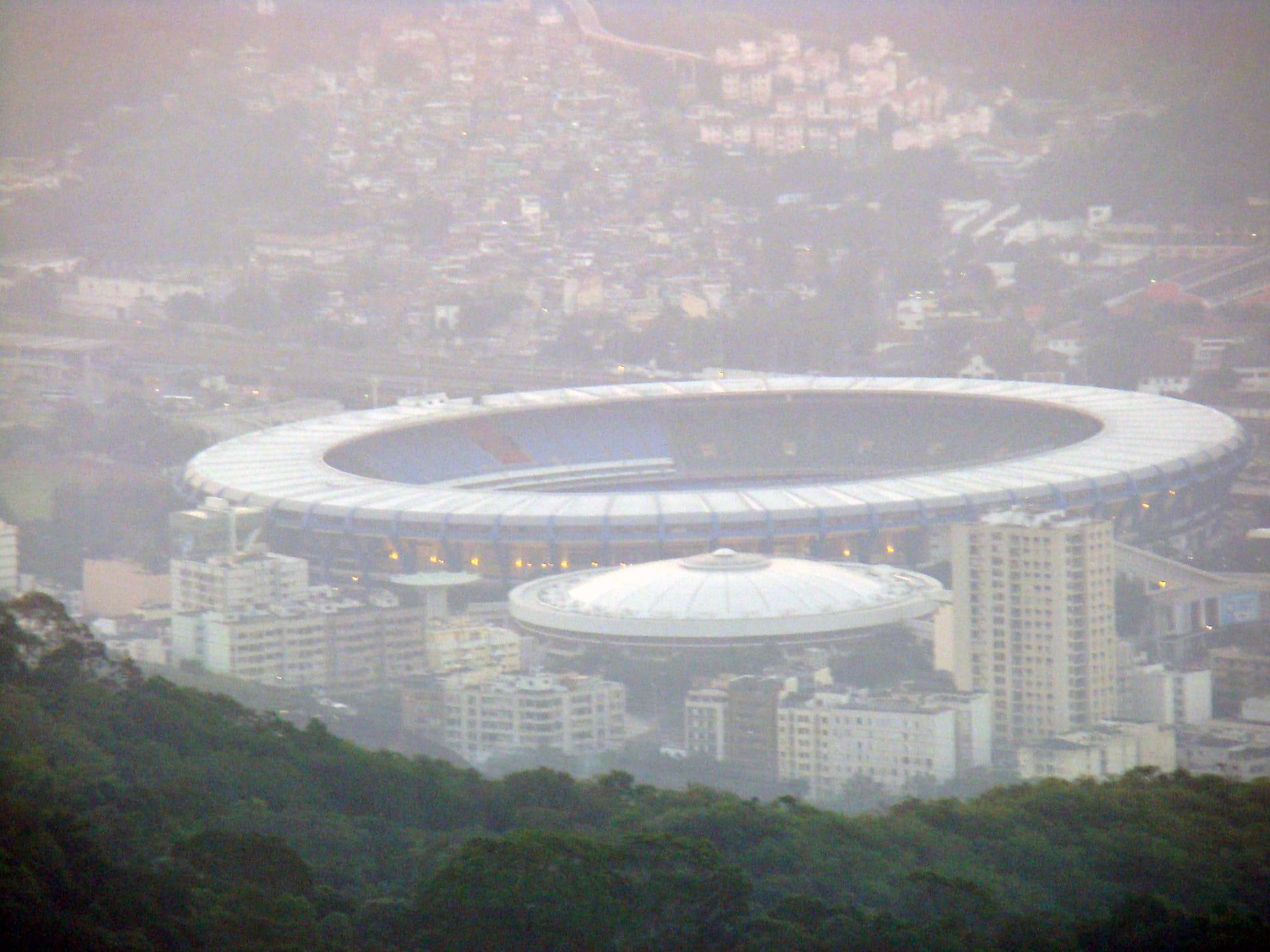 Rio de Janeiro, Brasil. O Estádio Jornalista Mário Filho, mais conhecido como Maracanã, é um estádio de futebol localizado no Rio de Janeiro e inaugurado em 1950, tendo sido utilizado na Copa do Mundo de Futebol daquele ano. Desde então, o Maracanã foi palco de grandes momentos do futebol brasileiro e mundial, como o milésimo gol de Pelé, finais do Campeonato Brasileiro e Carioca de Futebol, competições internacionais e partidas da Seleção Brasileira. Foi um dos locais de competição dos Jogos Pan-Americanos de 2007, recebendo o futebol, as cerimônias de abertura e de encerramento. Ao longo do tempo, no entanto, o estádio também passou a assumir caráter de espaço multiuso ao receber outros eventos como espetáculos e partidas de outros esportes, como o voleibol em uma oportunidade. Após diversas obras de modernização, a capacidade atual do estádio é de 92 mil espectadores,[3] sendo o maior estádio do Brasil. O nome oficial do estádio, Mário Filho, foi dado em homenagem ao falecido jornalista carioca, irmão de Nelson Rodrigues, que se destacou no apoio à construção do Maracanã. Já o nome popular é oriundo do rio Maracanã, que cruzava a Tijuca e São Cristóvão, desaguando na baía de Guanabara. Em tupi-guarani, a palavra Maracanã significa "semelhante a um chocalho".[25] Devido à construção do estádio, foi criado o bairro do Maracanã, onde o estádio fica localizado, originalmente parte da Tijuca. Nesta área da cidade, existiam diversas aves, vindas da Região Norte do Brasil, conhecidas como Maracanã-guaçu. ________________________________________________________________ The Estádio Jornalista Mário Filho, commonly called Estádio do Maracanã ("Maracanã stadium"), is an open-air stadium in Rio de Janeiro, Brazil. Owned by the Rio de Janeiro State Government, it is named after the Maracanã neighbourhood in Rio de Janeiro. It was opened in 1950 to host the FIFA World Cup. Since then, it has mainly been used for football matches between the major football clubs in Rio de Janeiro, including Flamengo, Fluminense, Botafogo and Vasco da Gama. It has also hosted a number of concerts and other sporting events. Although the paid attendance at the final game of the 1950 FIFA World Cup was 199,854, the stadium currently seats 88,992 spectators.[1] Despite the reduction in capacity, it remains the second largest stadium in South America behind Estadio Monumental Banco Pichincha in Guayaquil, Ecuador. It is due to host the World Cup Final in the 2014 World Cup, becoming the second stadium to host football's most important match twice after the Azteca Stadium in Mexico City.