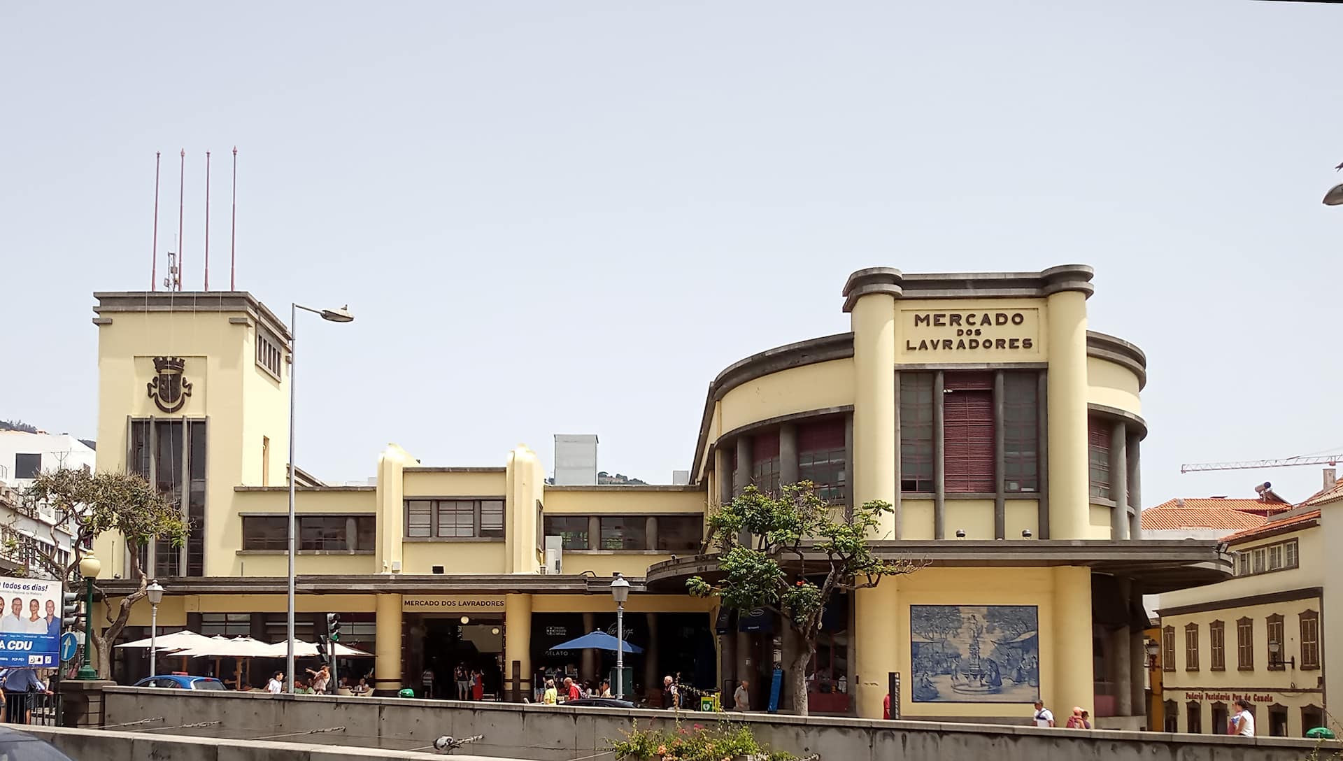 Mercado dos Lavradores Edmundo Tavares (1892-1983), 1938 a 1940." O Mercado dos Lavradores foi projetado pelo arquiteto Edmundo Tavares (Oeiras, 8 nov. 1892-Lisboa, 9 abr. 1983) em 1938 e inaugurado a 24 nov. 1940, pelo Dr. Augusto da Silva Branco Camacho (1907-1991), natural da Ponta do Sol, servindo de governador civil, na vigência da presidência da câmara do Funchal do Dr. Fernão Ornelas (1908-1978), presidente da Comissão Administrativa da Câmara Municipal do Funchal de 12 jan. 1935 a 22 out. 1946, encontrando-se presente o presidente da Junta Geral, Dr. João Abel de Freitas (1893-1948), o tenente-coronel Francisco Silvestre Varela (1883-1969), governador militar e o cónego magistral Manuel Francisco Camacho (1877-1970). Fotografia de 10 de agosto de 2023. Rua Brigadeiro Oudinot, Funchal, ilha da Madeira O Projeto de Melhoramentos para o Funchal de Edmundo Tavares 1933-1936 (c.) Duas plantas nos arquivos da Câmara Municipal, uma das quais assinada e com o título supracitado, mas não datadas, parecem indiciar o primeiro trabalho para a edilidade funchalense deste arquiteto, depois autor de dois dos mais icónicos edifícios da cidade: o Banco de Portugal (1934-1940) e o Mercado dos Lavradores (1938-1940), este, provavelmente, também um dos mais conseguidos da sua época. A confirmar-se a autoria da planta não assinada nem datada, deve a mesma datar de algum tempo depois da estadia do arquiteto Carlos Ramos (1897-1969) no Funchal, em março de 1932, quando se definiu com alguma segurança o traçado da futura Avenida do Infante, então somente equacionada como a via principal do “ Bairro dos Ilhéus ”, e que esse arquiteto propõe que fosse dotada de habitações unifamiliares e independentes, obedecendo a uma única gramática arquitetónica “ à antiga portuguesa ”, a “ uma arquitetura tradicionalista, com os seus beirados, azulejos, rótulas, pináculos, vasos de flores, etc., etc .”, algumas das quais habitações seriam depois projetadas por Edmundo Tavares (1892-1983), único arquiteto com residência na Ilha nestes anos. A planta em causa define assim o traçado da futura Avenida do Infante, assim como a sua sequência para a Avenida Arriaga, que necessitaria de ser alargada para Norte, em parte à custa de uma faixa do Jardim Municipal, o que será efetuado em 1942. Aponta-se, igualmente, a futura intervenção no palácio Carvalhal-Albuquerque, sede da edilidade e que será depois projetada numa parceria de Carlos Ramos com Raul Lino (1879-1974), obra que vai decorrer em 1942, indicando-se a necessidade de aproveitamento da então cerca do Liceu do Funchal, instalado no antigo Paço Episcopal, para a constituição de um futuro largo frente à sede camarária. Nesta planta aparece também a indicação de haver intervenção na área da foz das ribeiras de Santa Luzia e de João Gomes, devendo estar em equação a demolição do então mercado D. Pedro V e a construção de uma ponte sobre essa foz, depois integrante da futura Avenida do Mar. A planta dos melhoramentos de Edmundo Tavares apresenta a planta do Liceu do Funchal numa versão muito inicial, provavelmente, de 1936, articulando-se para nascente com o Mercado Municipal, numa configuração e numa localização depois abandonadas. O mercado veio a ser construído bastante mais para poente do liceu, adaptando-se ao quarteirão das ruas Latino Coelho e do Hospital Velho, tendo o seu projeto final sido aprovado em 1938, mas já estando perfeitamente equacionado em meados de 1937 (Cf. Re-Nhau-Nhau , Funchal, 28 jun. 1937, p. 10). O arquiteto Edmundo Tavares nasceu em Oeiras, a 8 nov. 1892 e faleceu em Lisboa, a 9 abr. 1983. Arquiteto e professor, foi discípulo de José Luís Monteiro (1848-1942) e fez o curso da Escola de Belas Artes de Lisboa entre 1903 e 1913, com uma das mais altas classificações de todos os cursos lecionados naquela escola nesses anos. Em 1914 já era arquiteto da Câmara de Lisboa, assinando as plantas e alçados da ampliação do Mercado 24 de Julho (Mercado da Ribeira de Lisboa) e, em 1915, expôs na SNBA uma série de projetos de Casas de Estilização Portuguesa , tendo sido premiado com várias medalhas de bronze e prata. Pouco depois de terminado o curso, em 1913, foi quarto premiado, com o escultor Maximiano Alves (1888-1954), no concurso do monumento ao Marquês de Pombal ( Ilustração Portuguesa , 27 abr. 2014, p. 256), tal como em 1916 seria também premiado nos concursos para os edifícios dos Paços do Concelho de Guimarães e do Porto. Seria ainda galardoado com o prémio do concurso de pensionistas arquitetos no estrangeiro do legado Valmor , em Paris (1919) e em Roma (1922); terceiro premiado no concurso para a construção do Liceu Dr. Júlio Henriques, em Coimbra (1930), etc. Em 1920 fez para a Madeira, Santa Maria Maior, o projeto do palacete do juiz Barata Monteiro, trabalho ao gosto Fim de Século , concorrendo, entretanto ao lugar de professor da Escola Industrial e Comercial do Funchal, cidade onde estava em finais de 1932 e de onde saiu em 1939, para idêntico lugar na Figueira da Foz. Trabalhando sempre paralelamente na atividade liberal, alguns dos seus projetos do Funchal encontram-se fora das datas da sua estadia na Madeira. Assinou, assim, uma vasta obra notável dentro da linguagem modernista e dos cânones da Art Deco , por vezes apelidada de Brutalista e, alternadamente e em oposição, dentro dos cânones do Português Suave , que equacionara já na sua exposição de 1915, na SNBA, em Lisboa e que voltará a desenvolver e teorizar no Funchal, ilustrando e colaborando com o major João dos Reis Gomes (1869-1950) nas Casas Madeirenses (1937), como o mesmo escreve nessa edição, que havia anos que vinham abordando esse tema, assunto a que este arquiteto voltará em Lisboa, em A Habitação Portuguesa, Casas Modernas, Vivendas Portuguesas (1952). Para além de algumas dezenas de habitações, algumas na icónica Avenida do Infante, projetou também unidades hoteleiras, de saúde e educacionais, inclusivamente, uma capela, devendo poucos arquitetos na Madeira terem deixado uma obra desta envergadura e, para mais, no curto espaço de tempo de sete anos. Como na abertura desta entrada assinalámos, o projeto da agência do Banco de Portugal (1934), de linguagem eclética e historicista, de fachada cenográfica neobarroca e profusamente ornamentada, parcialmente reforçada com cantaria cinzenta local e coberto por alto telhado piramidal sextavado, encimado por esfera armilar, tornou-se dos mais icónicos edifícios da cidade, estabelecendo, de certa forma, diálogo com a torre da Sé, tal como com a estátua de Zarco (1928-1934), de Francisco Franco (1885-1955). Interiormente, ainda reforça a cenografia neobarroca, mesmo, quase religiosa, através da iluminação do átrio central por claraboia, seguindo o esquema de articulação interior do antigo Banco Nacional Ultramarino (1924-1926), no seguinte quarteirão, edifício igualmente dotado de cobertura piramidal, mas mais discreta, pois que algo recuada em relação à fachada, é pouco percetível pelo transeunte. O seu mais icónico edifício, no entanto, deve ser o do Mercado dos Lavradores (1938-1940), com toda uma outra linguagem muito mais arejada, em alvenaria mista com frisos relevados de betão aparente e de tijoleira, que igualmente reforçam o embasamento, tal como dotado de amplos painéis de azulejos com temas regionais em azul e branco, com molduras policromadas, trabalhos da Faiança Battistini de Maria Portugal (Lepoldo Battistini, 1865-1936), assinados e datados de 1940. O edifício envolve um amplo átrio central, a céu aberto e é percorrido lateralmente por terraços, igualmente abertos, integrando para nascente uma praça de peixe. Não deixando de ser um edifício Estado Novo e dito Português Suave , é assim e em principio, uma das obras mais bem conseguidas do seu tempo. Rui Carita Universidade da Madeira. Bibliografia: FRANCO, Sérgio Miguel Gouveia (2012), A Obra de Edmundo Tavares no Funchal , Universidade Fernando Pessoa, Faculdade de Ciências e Tecnologia, Curso de Arquitetura e Urbanismo, Porto; GOMES, João dos Reis (1937), Casas Madeirenses , colaboração de Edmundo Tavares, Funchal, Diário da Madeira ; Ilustração Portuguesa (1914), n.º 427, “Projeto do Monumento ao Marquês de Pombal”, Lisboa, 27 abr. 1914, p. 526; TAVARES, Arq. Edmundo (1952), A Habitação Portuguesa: Casas Modernas, "'Vivendas Portuguesas , Bertrand Irmãos; Ib. (1980), A madeira na construção civil, seleção de trechos de carpintaria: portas, janelas e acessórios , 2ª edição, Silvas, cop. Trab. Graf. Scael, Lisboa; Re-Nhau-Nhau (1937), “A Câmara Obra!”, Funchal, 28 jun. 1936, p. 10; VASCONCELOS, Teresa (2008), O Plano Ventura Terra e a Modernização do Funchal (Primeira Metade do século XX) , tese de mestrado, Coleção Funchal 500 Anos, n.º 5.