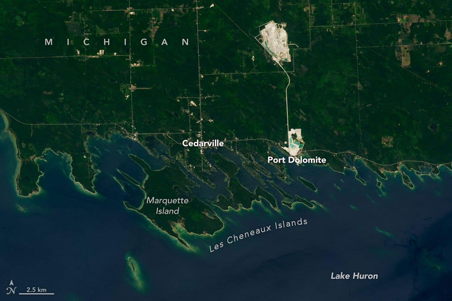 A portion of the Upper Peninsula of Michigan runs across the top of the image. It is mostly green, with some roads and two bright rock quarries visible. Numerous islands near the shore have elongated shapes and are oriented at a diagonal.