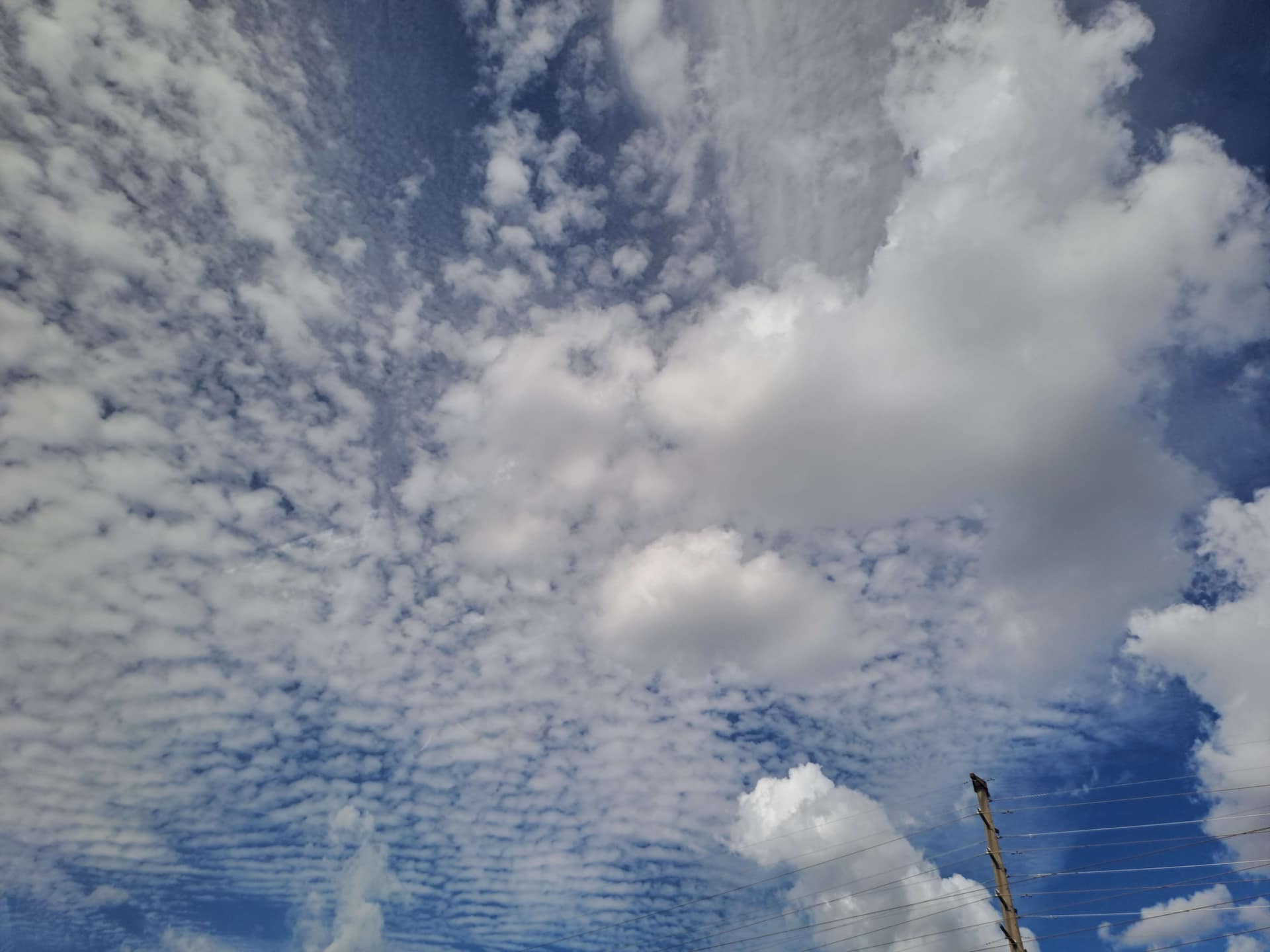 Céu azul coberto com nuvens de diversos formatos. Um poste fica no canto inferior esquerdo.